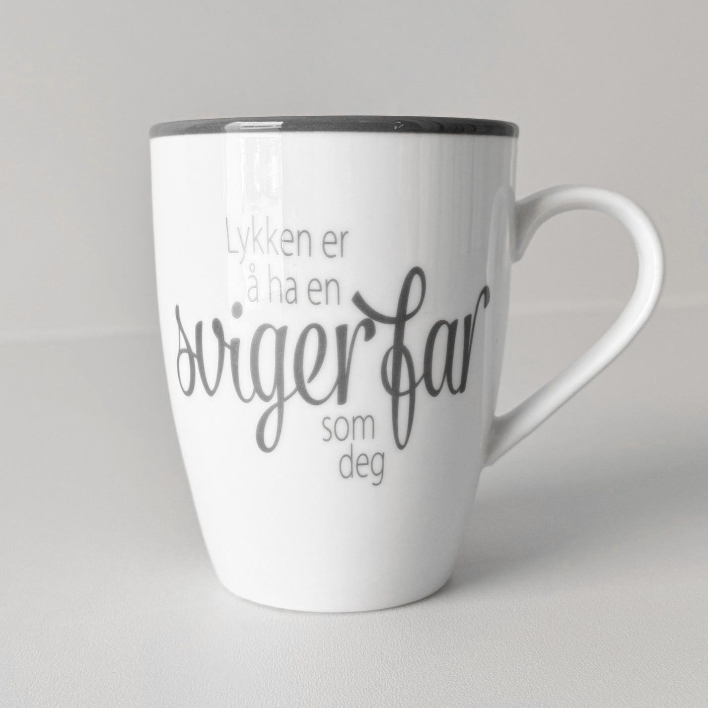 Hvit keramikkopp med teksten "Lykken er å ha en svigerfar som deg" mot lys, nøytral bakgrunn.