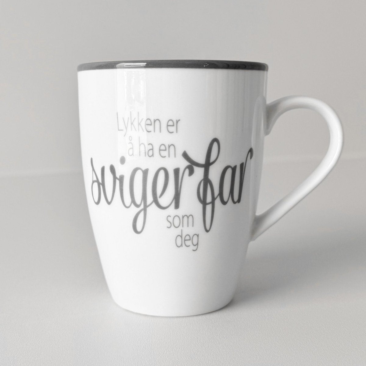 Hvit keramikkopp med teksten "Lykken er å ha en svigerfar som deg" mot lys, nøytral bakgrunn.