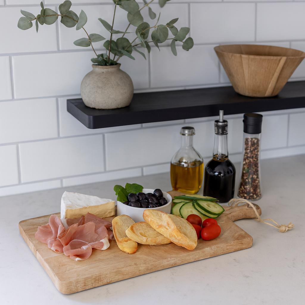 Rustikk skjærefjøl på kjøkkenbenk, fylt med ost, skinke, brød, oliven, tomater og agurk, med olje og krydder i bakgrunnen.