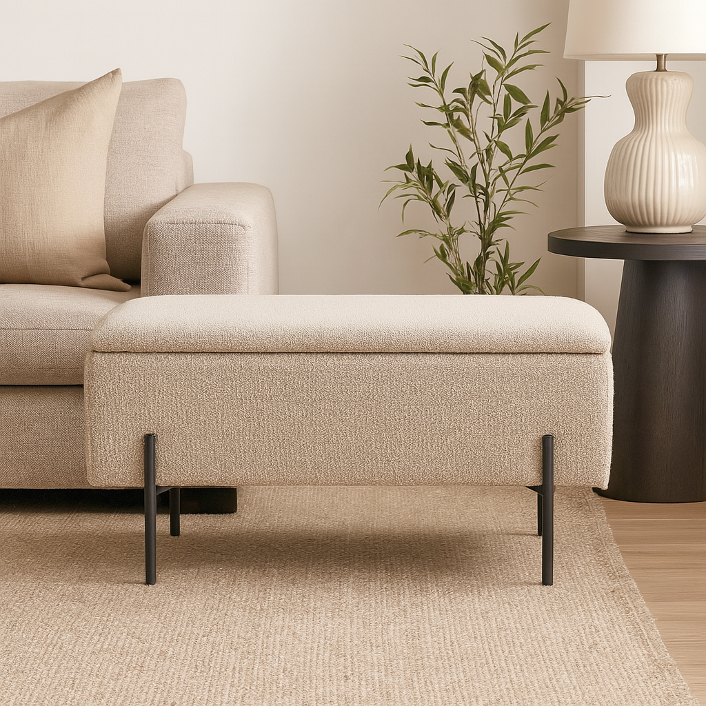 Elegant polstret benk i beige stoff med slanke svarte ben, stående på et teppe foran en sofa omgitt av et bord og grønn plante.