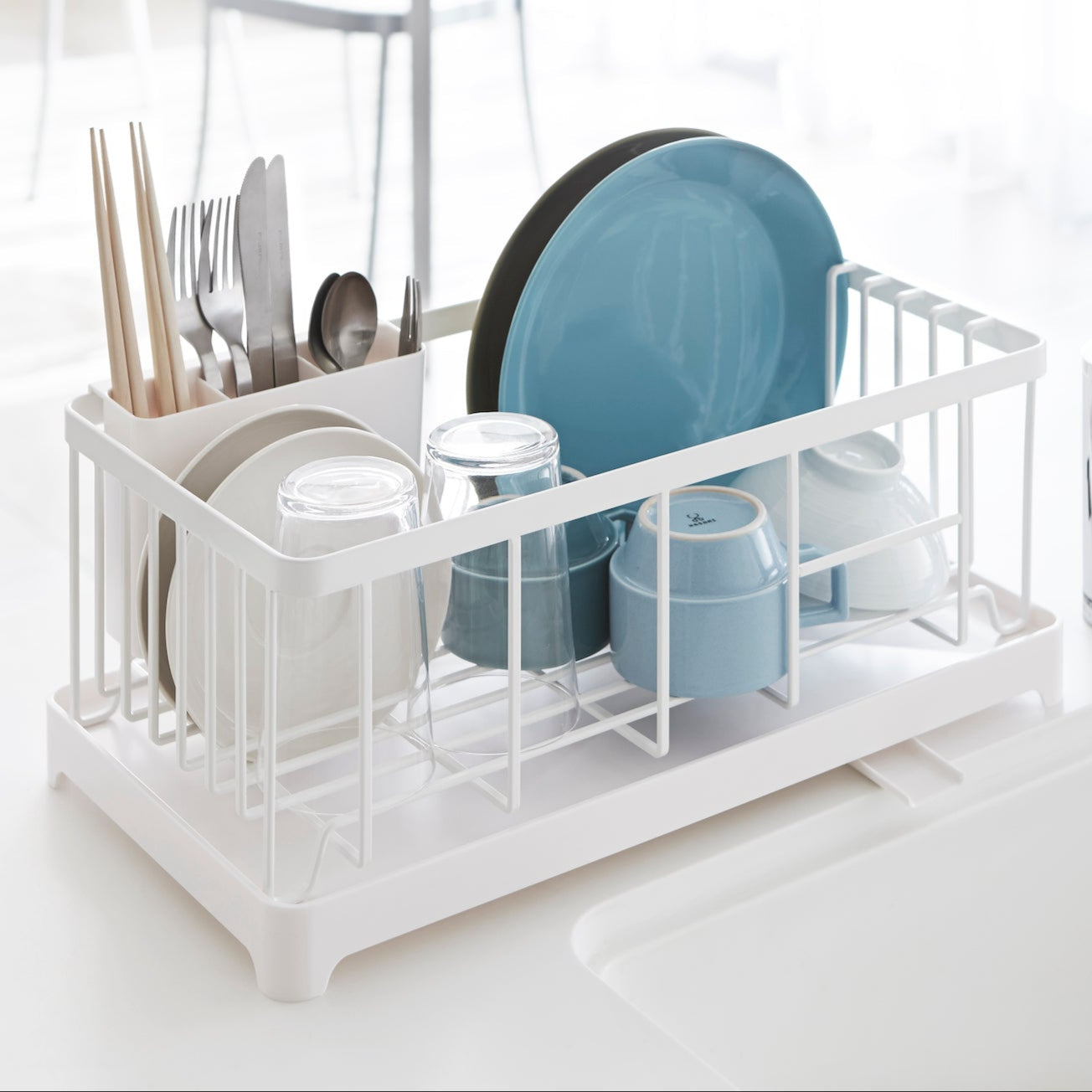 Hvit oppvaskstativ med drypprenne plassert ved vasken, fylt med tallerkener og glass.