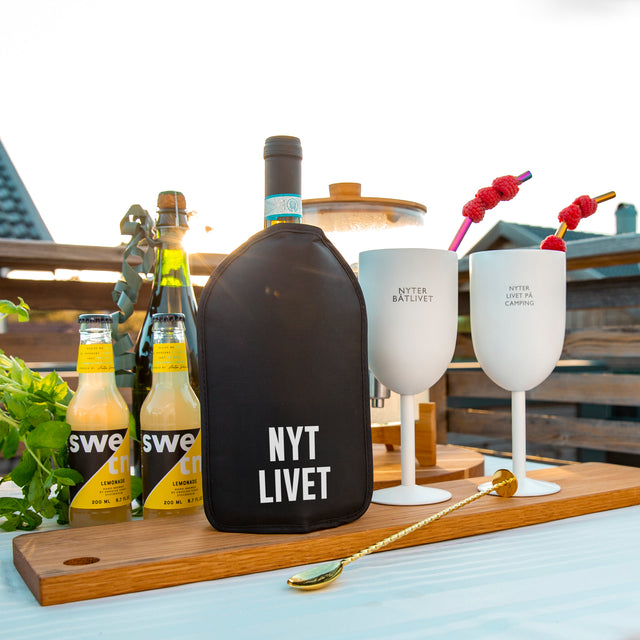 Sort vinkjøler med teksten Nyt Livet, plassert på bord med drikke og glass.