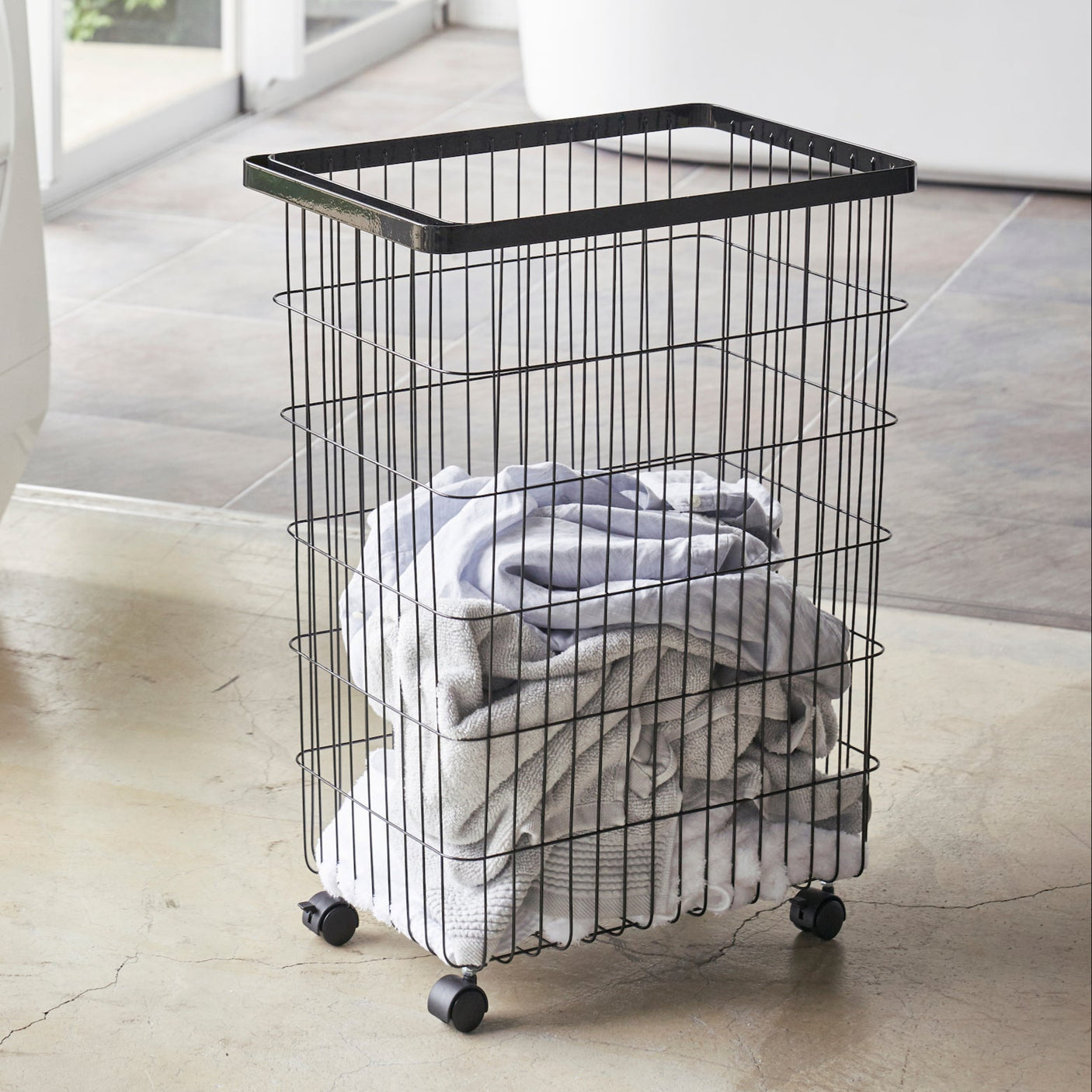 Sort skittentøyskurv på hjul i metall, fylt med håndklær, plassert på vaskeromsgulv.