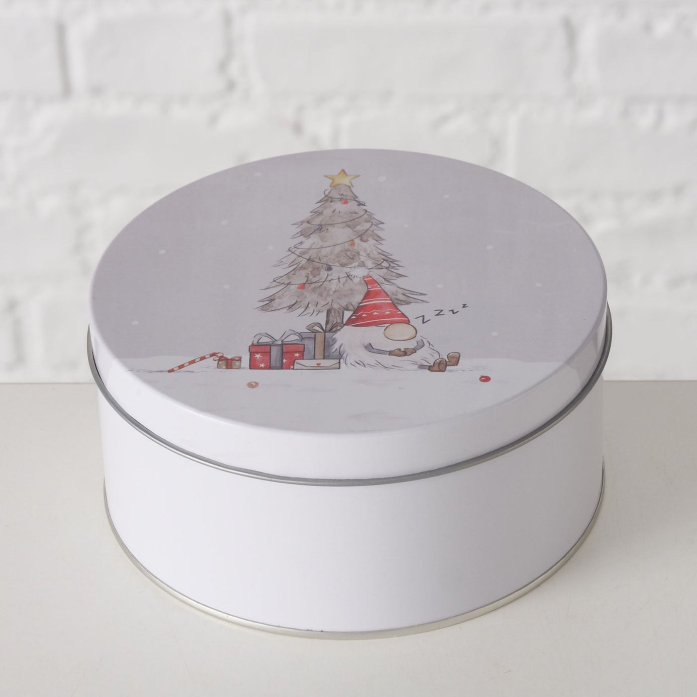 Hvit kakeboks med julemotiv av sovende nisse ved juletre og gaver, mot lys mursteinsvegg.