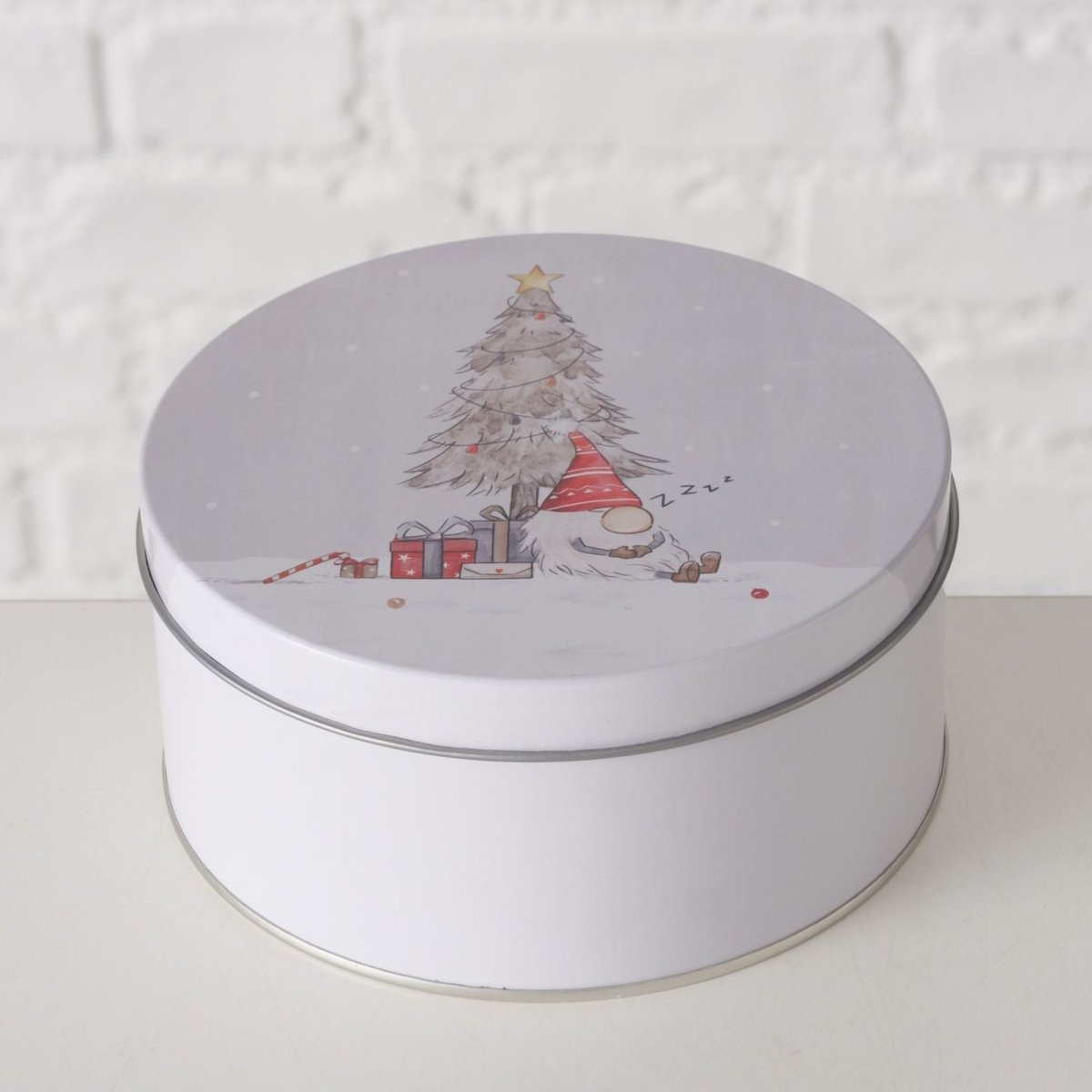 Vit kakorburk med julmotiv av en sovande tomte vid juleträ och gåvor, mot en ljus mursteinvägg