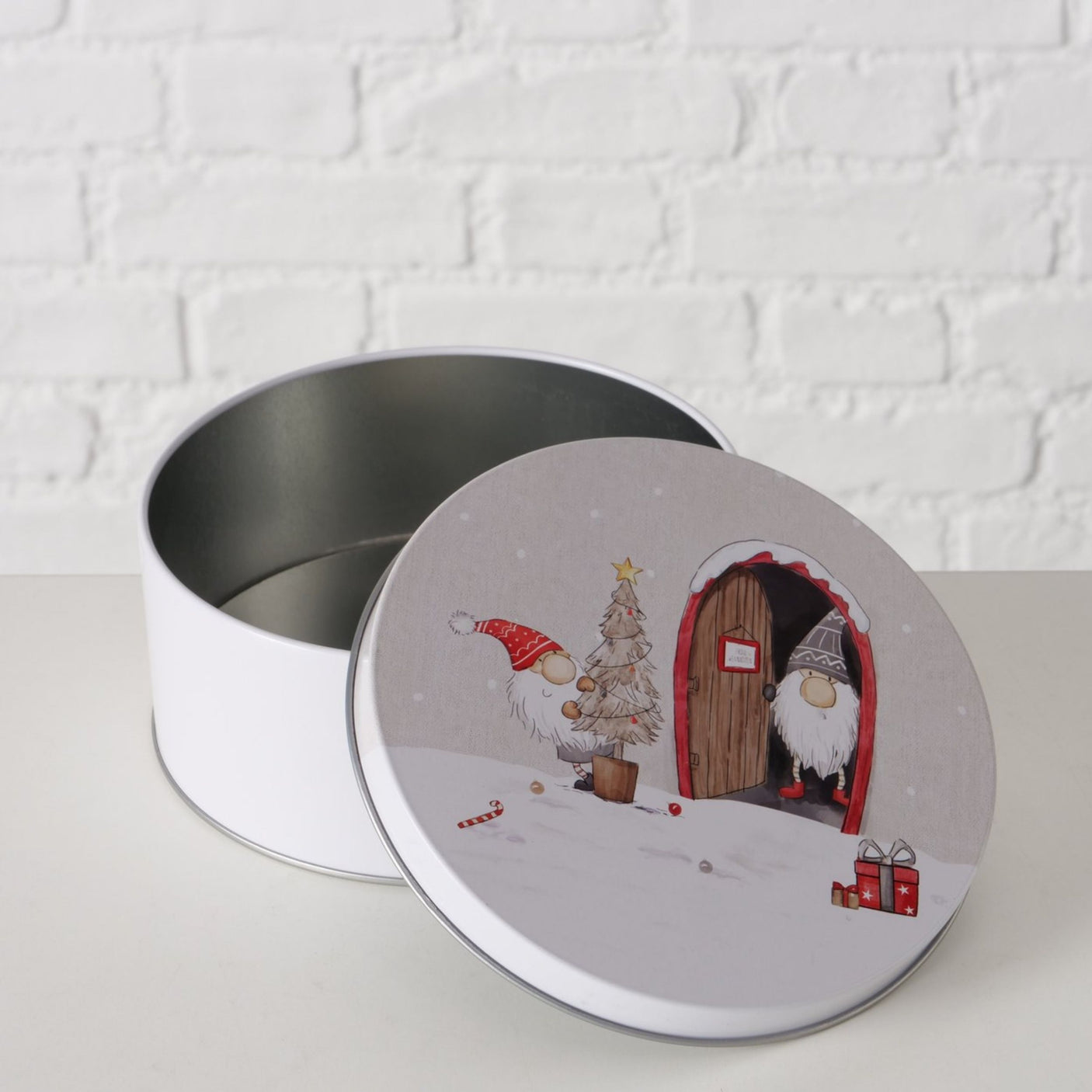 Hvit kakeboks med julemotiv av to nisser, én ved dør og én med tre, mot lys mursteinsvegg.