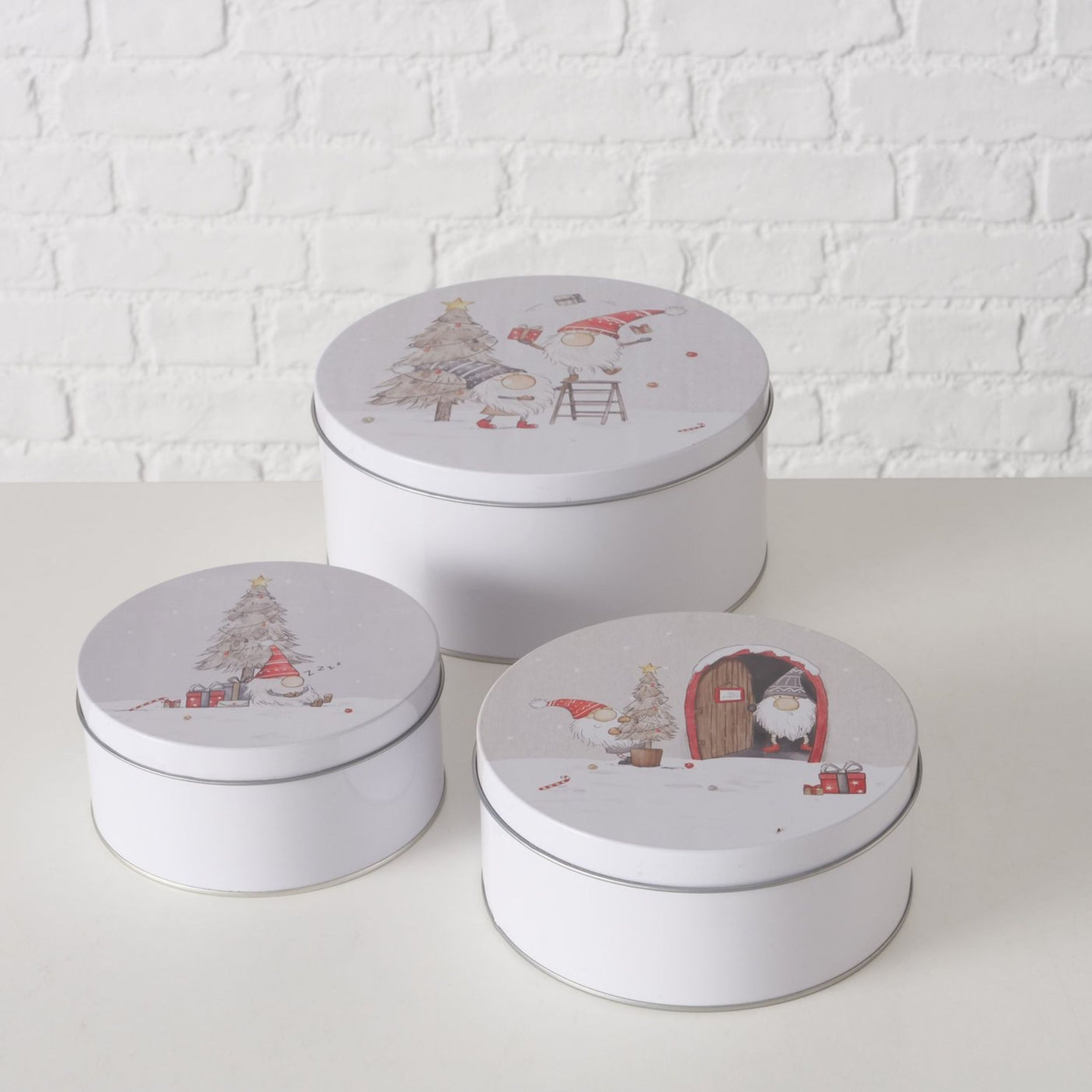 Tre hvite kakebokser med julemotiv av nisser og juletre mot lys mursteinsvegg.