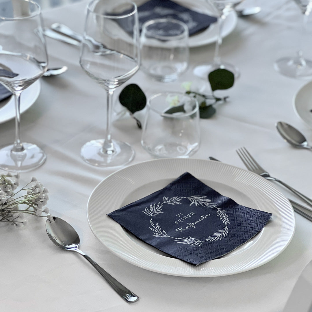 Mørkeblå serviett med teksten «Vi feirer konfirmanten» ligger på tallerken i elegant borddekking.