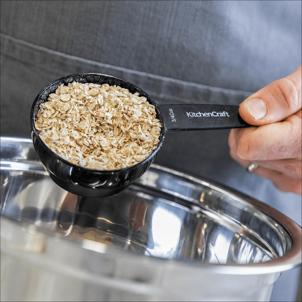 Svart KitchenCraft-mätkopp fylld med havregryn, hållen över en rostfri stålbolle.