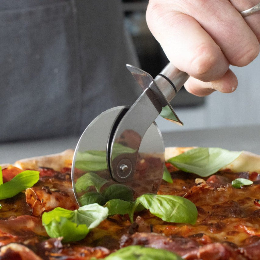 Hånd bruker et pizzahjul i rustfritt stål til å skjære en pizza med ost, tomat og frisk basilikum. - Lindas dekor