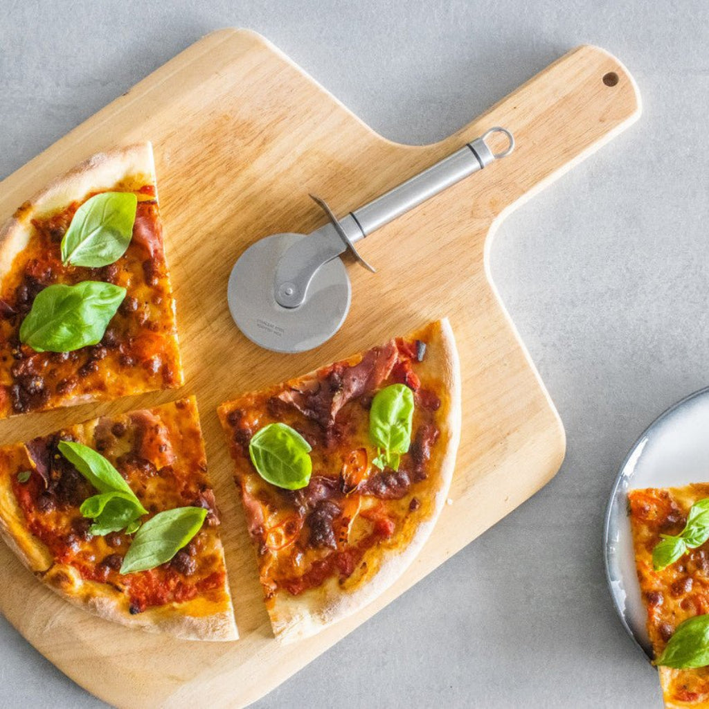 Pizza skåret i stykker på en trefjøl med pizzahjul i rustfritt stål liggende ved siden av. - Lindas dekor