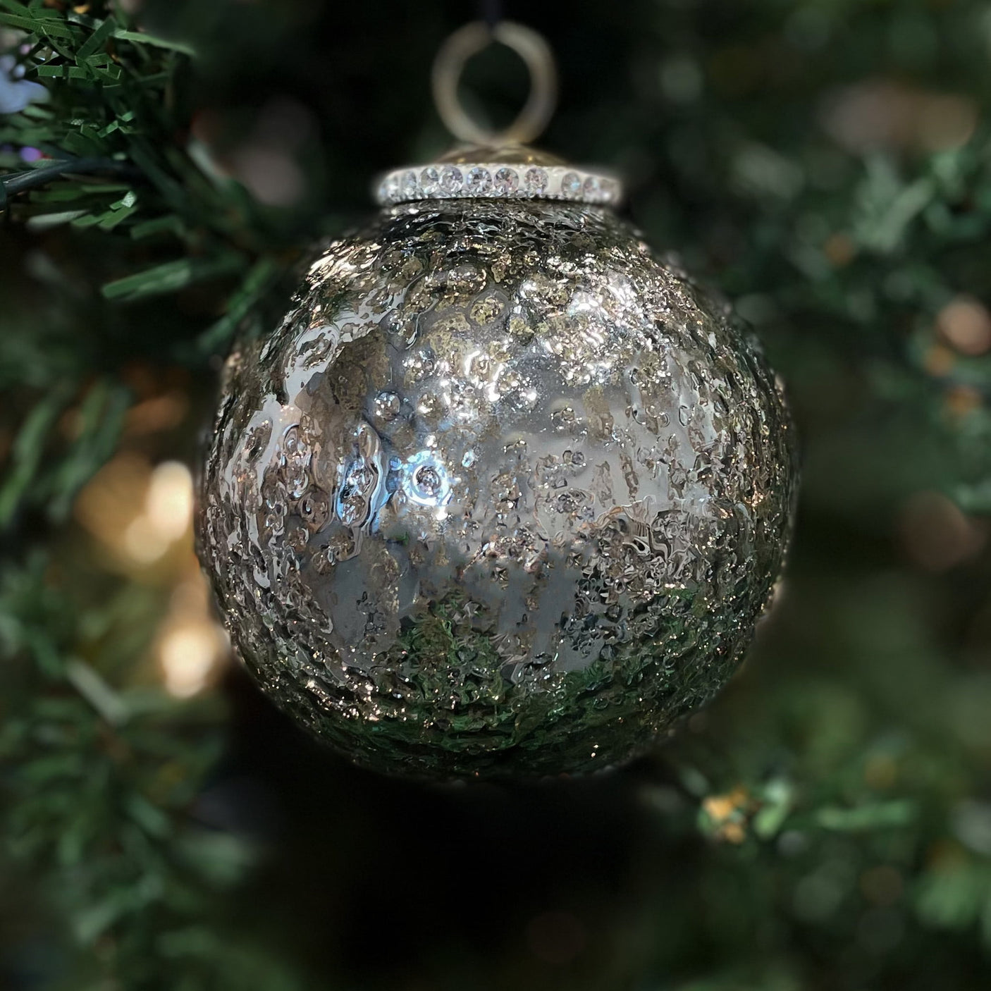 Grå julekule i glass – henger på juletre med grønne greiner og julelys i bakgrunnen.