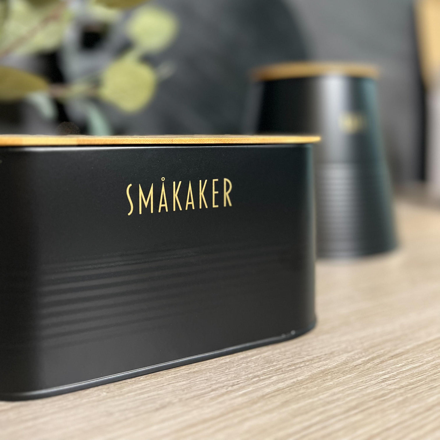 Elegant småkakeboks i sort metall med bambuslokk og gulltekst, plassert på kjøkkenbenk.