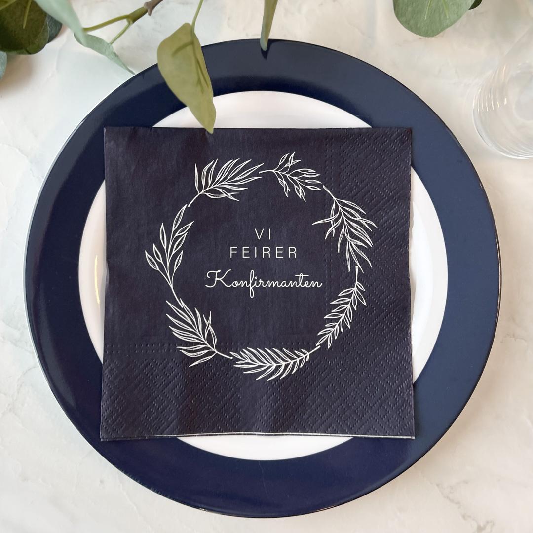 Nærbilde av mørkeblå serviett med hvit krans og tekst på tallerken.