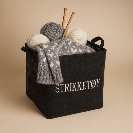 Sort stoffkurv med tekst "STRIKKETØY", fylt med garnnøster, halvstrikket genser og strikkepinner.