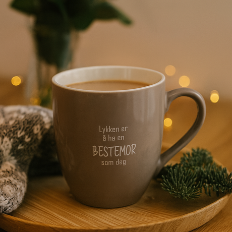 Beige kopp med teksten "Lykken er å ha en BESTEMOR som deg" på trebord med julelys og granbar.