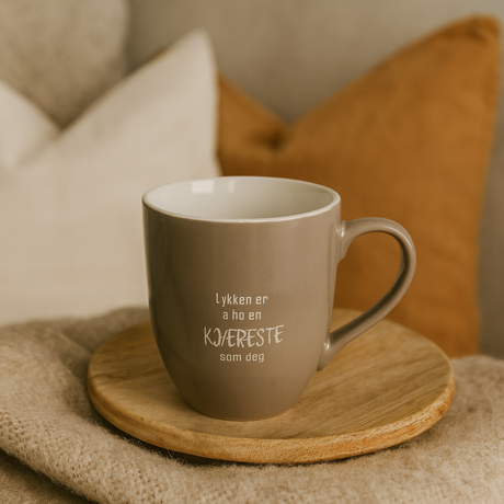 Beige kopp med teksten «Lykken er å ha en kjæreste som deg» på brett, foran puter i varme toner.