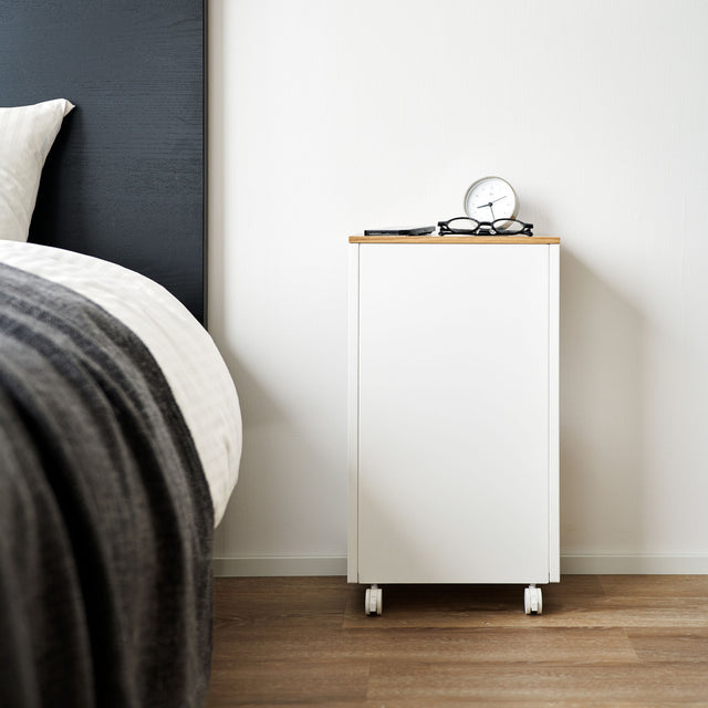 Hvit sidebord med treplate og hjul plassert ved seng med lampe og sengeteppe.