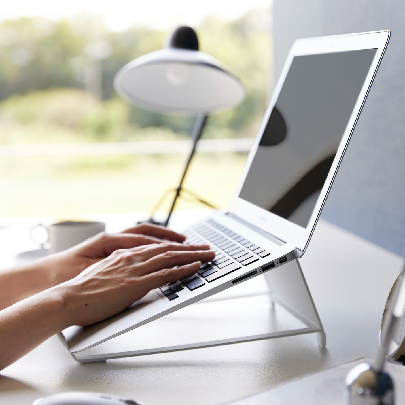 Hvit justerbar laptopstativ brukt på skrivebord ved vindu.