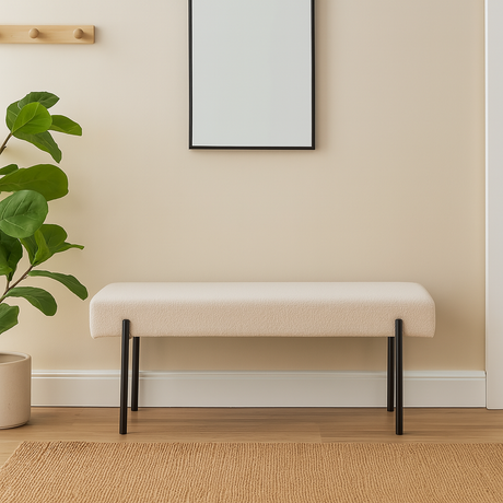 Beige benk med svarte metallben står mot en lys vegg i en pyntet gang med grønn plante, veggkroker og lys tremønstret gulv med vevd teppe.