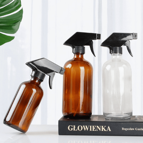 Tre sprayflasker i glass, to i brunt og én i klart glass, med svarte spraytopper, plassert dekorativt på en bok.