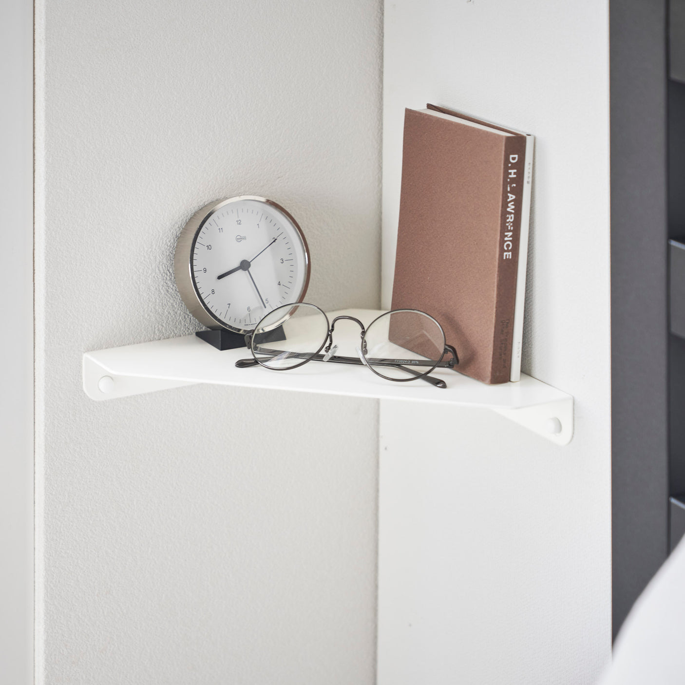 Minimalistisk hvit hjørnehylle med klokke, briller og bok plassert som dekor og oppbevaring.