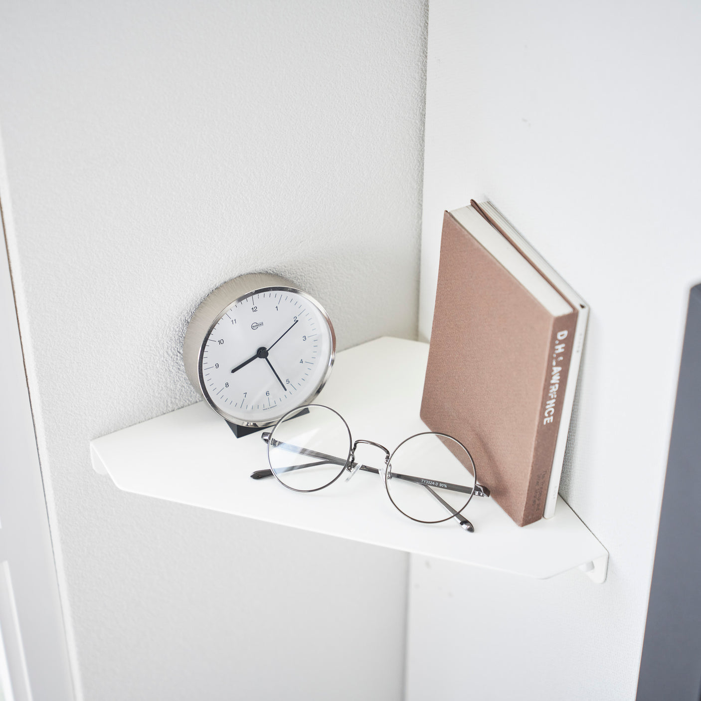 Nærbilde av hvit hjørnehylle med klokke, bok og briller arrangert pent på overflaten.
