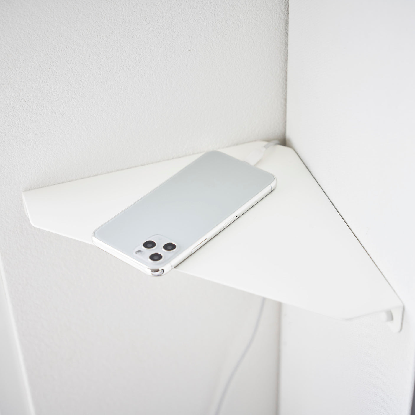 Hvit vegghylle montert i hjørnet, brukt som ladestasjon for en mobiltelefon som ligger på hyllen.