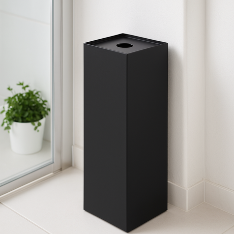 Sort dorullholder i lukket modell med synlig hullåpning på toppen, plassert ved siden av dusjdør på et lyst moderne bad. Minimalistisk design som gir praktisk og plassbesparende oppbevaring av toalettpapir.
