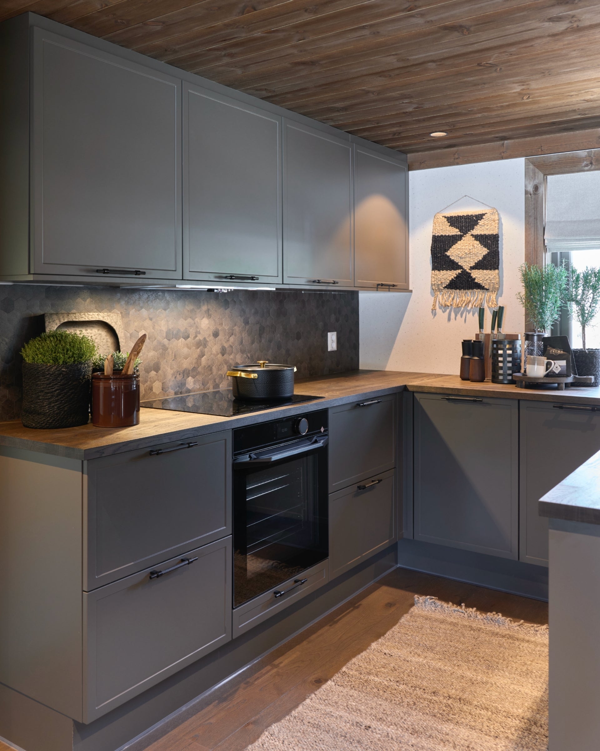 Moderne hyttekjøkken i Setesdal med mørkebrunt tretak, grå fronter, og Hexagon mørk betong selvklebende fliser fra Lindas Dekor som backsplash bak komfyr og benkeplate.