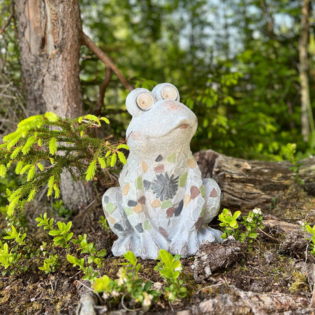 En dekorativ froskefigur står i skogbunn omgitt av mose og små grønne planter, med trær i bakgrunnen.