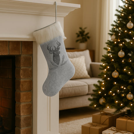 Grå julestrømpe i filt med reinsdyrmotiv og hvit pelskant, hengende på peis ved juletre pyntet med lys og julekuler.