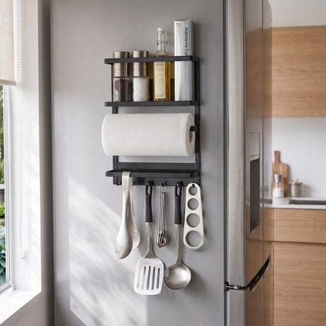 Sort magnetisk hylde monteret på siden af et køleskab, organiseret med krydder, olje, kjøkkenpapir og køkkenredskaber i et lyst, moderne køkken.