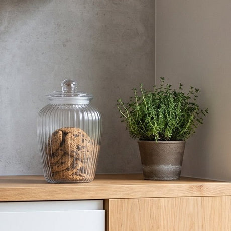 Lyst, moderne kjøkken med hvite fronter og detaljer i tre, dekorert med en riflet glasskrukke fylt med kjeks, svart vannkoker og en grønn urteplante på kjøkkenbenken.